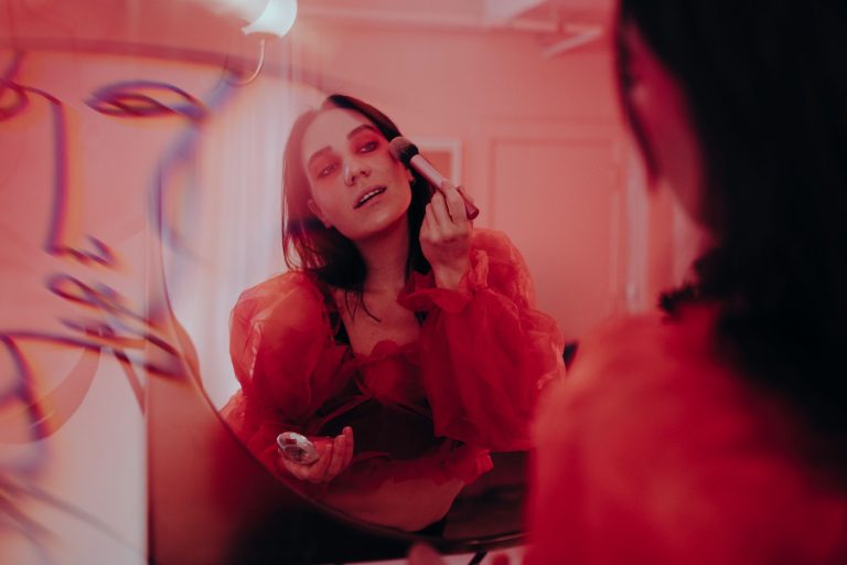 A woman applies makeup in front of a mirror reflecting an artistic red ambiance, creating a stylish, creative scene. — Moistu